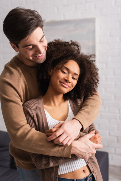 Positive man hugging african american girlfriend at home  - Photo, Image