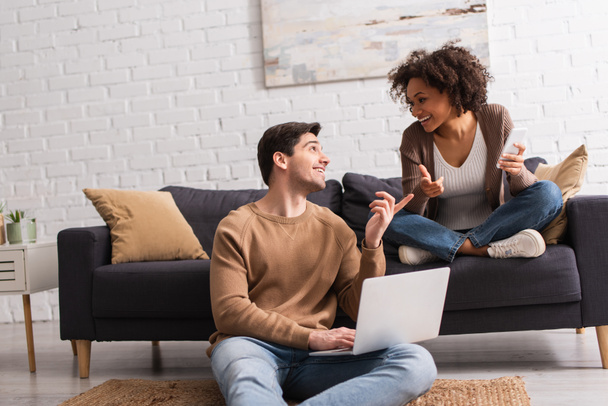 Sorrindo casal interracial com dispositivos apontando com os dedos em casa  - Foto, Imagem