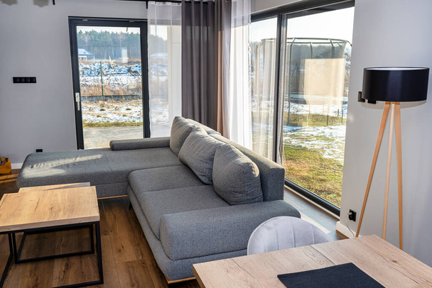 Uma moderna e simples sala de estar na casa com um sofá cinza, grandes janelas do pátio e mesas. - Foto, Imagem