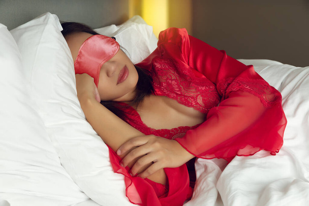 Femme asiatique en satin rouge vêtements de nuit avec masque pour les yeux dormir sur le lit dans la chambre - Photo, image