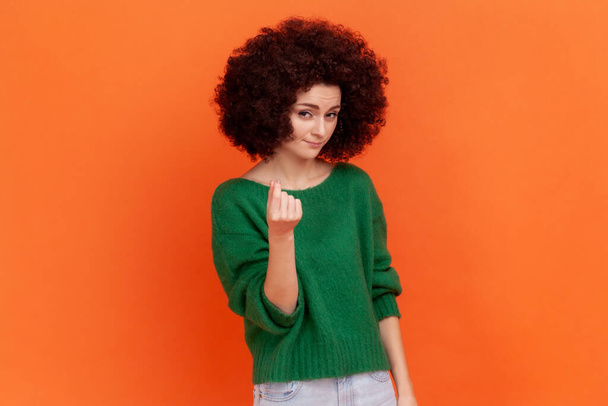 Woman with Afro hairstyle in green sweater rubbing fingers showing money gesture, asking for salary, demanding bribe, looking at camera with grin. Indoor studio shot isolated on orange background. - Foto, Bild