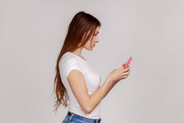 Side view of attractive serious woman using smart phone, typing message or checking social networks, writing post, wearing white T-shirt. Indoor studio shot isolated on gray background. - Foto, Imagen