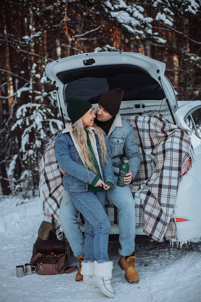 A loving young couple in an embrace sits in the trunk of a white car in winter. A young man in a brown hat, in a denim jacket and jeans. Young blonde girl in a green hat, green sweater, denim jacket and jeans. There are two hiking backpacks nearby. A - Zdjęcie, obraz