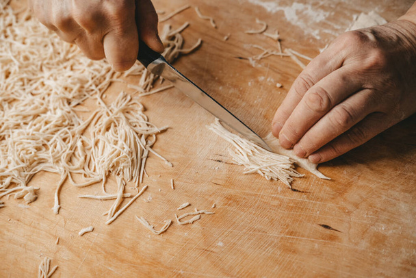 A woman cuts the dough with a knife and makes homemade noodles in the kitchen.Pasta homemade, by the hands of a woman. - 写真・画像