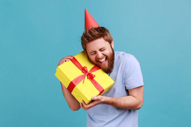 Portrait of happy positive handsome bearded man in party cone embracing yellow gift box, getting present from friend on his birthday. Indoor studio shot isolated on blue background. - Fotó, kép