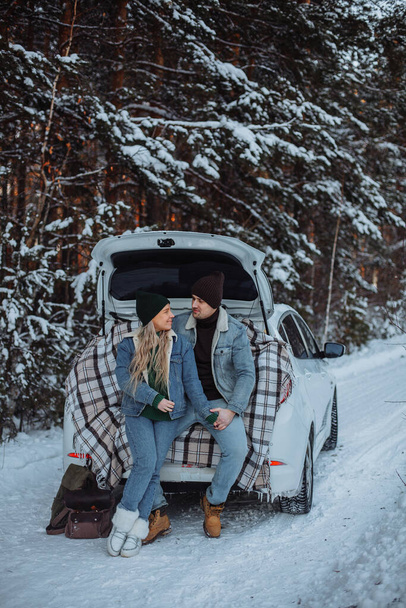 A loving young couple in an embrace sits in the trunk of a white car in winter. A young man in a brown hat, in a denim jacket and jeans. Young blonde girl in a green hat, green sweater, denim jacket and jeans. There are two hiking backpacks nearby. A - Foto, Imagem