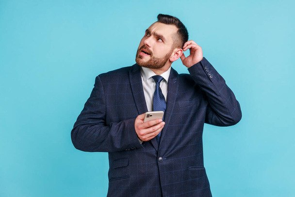 Businessman wearing official style suit scratching head holding smartphone in hand, contemplating about software updating, choosing suitable tariffs. Indoor studio shot isolated on blue background. - Foto, Imagen