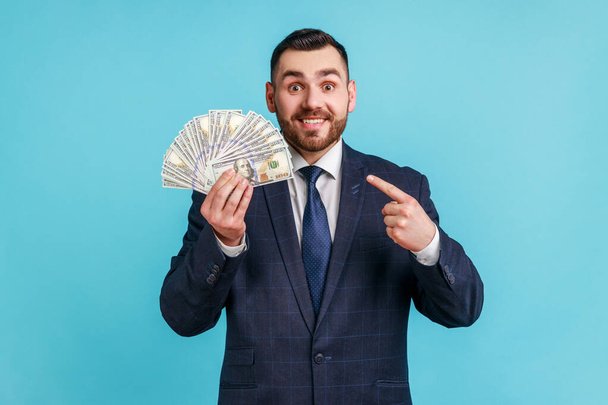 Businessman wearing official style suit holding lot of money, pointing at dollar bills in hand and smiling excitedly, enjoying lottery win. Indoor studio shot isolated on blue background. - Фото, изображение
