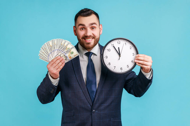 Portrait of happy businessman wearing official style suit showing dollar cash and wall clock on camera, hourly payment, looking at camera. Indoor studio shot isolated on blue background. - Foto, afbeelding