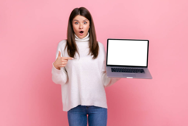 Shocked astonished female pointing at laptop display with blank white display for advertisement, wearing white casual style sweater. Indoor studio shot isolated on pink background. - Fotografie, Obrázek