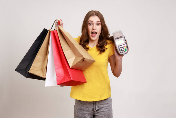 Shocked amazed female of young age in yellow T-shirt holding and showing payment terminal and shopping bags, expressing astonishment. Indoor studio shot isolated on gray background. - Foto, immagini