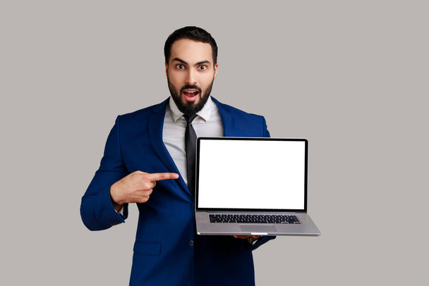 Shocked man pointing finger at laptop with blank display, showing empty screen, amazed with internet advertisement, wearing official style suit. Indoor studio shot isolated on gray background. - Photo, Image