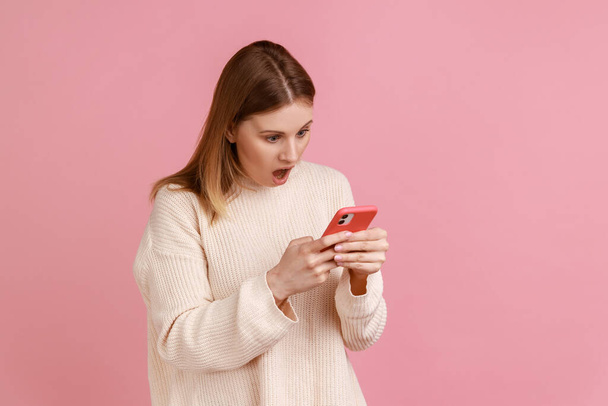 Portrait of beautiful astonished blond woman blogger using smart phone, looking at device screen, sees shocking content, wearing white sweater. Indoor studio shot isolated on pink background. - Photo, image