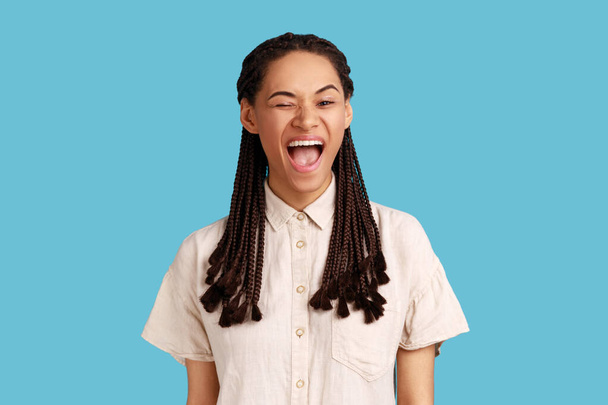 Portrait of good looking cheerful woman with black dreadlocks winking eye and flirts with someone, looks at camera, wearing white shirt. Indoor studio shot isolated on blue background. - Foto, Bild