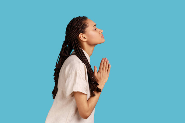 Side view of woman with dreadlocks keeps palms pressed together, pleading angel, has innocent expressions, asks for apologize, wearing white shirt. Indoor studio shot isolated on blue background. - Fotó, kép