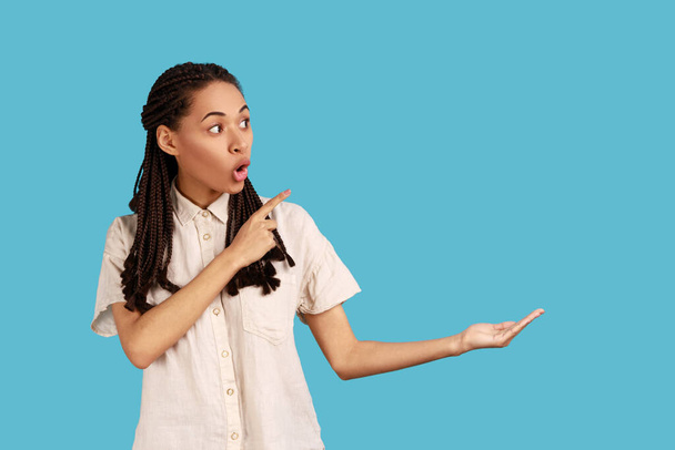 Look at commercial proposal. Shocked woman with dreadlocks pointing at copy space on her palm, empty place for idea presentation, product advertising. Indoor studio shot isolated on blue background. - Fotó, kép