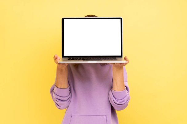 Portrait of unrecognizable woman hiding her face behind laptop with blank display for advertisement or promotional text, wearing purple hoodie. Indoor studio shot isolated on yellow background. - Photo, image