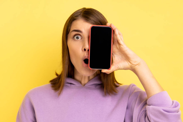 Attractive young adult woman covering eye with cell phone with empty display, having shocked expression, wearing purple hoodie. Indoor studio shot isolated on yellow background. - Foto, immagini