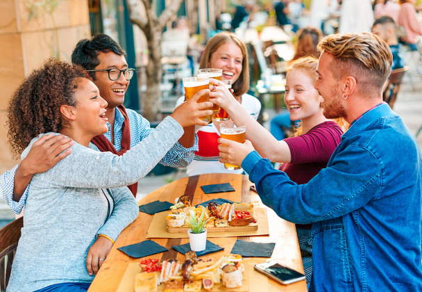 Tevreden multiraciale vrienden die aan tafel zitten en elkaar aankijken terwijl ze met bier op straat in de kantine staan - Foto, afbeelding