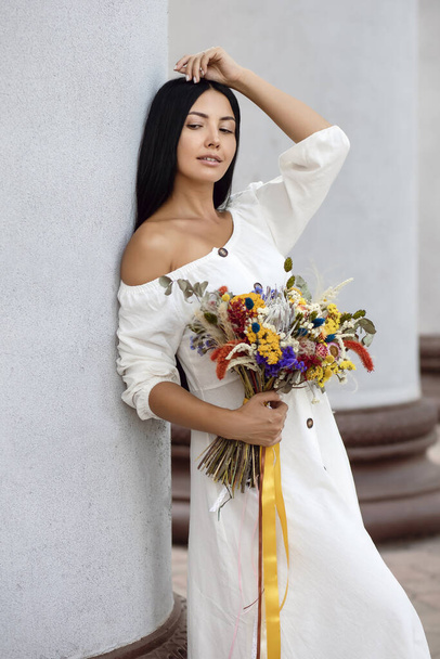 Eine schöne junge brünette Frau in einem weißen Kleid mit einem Strauß Wildblumen lehnte an einer Säule und posierte. Vertikales Porträt eines netten Mädchens mit langen schwarzen Haaren im Vintage oder rustikalen Stil - Foto, Bild