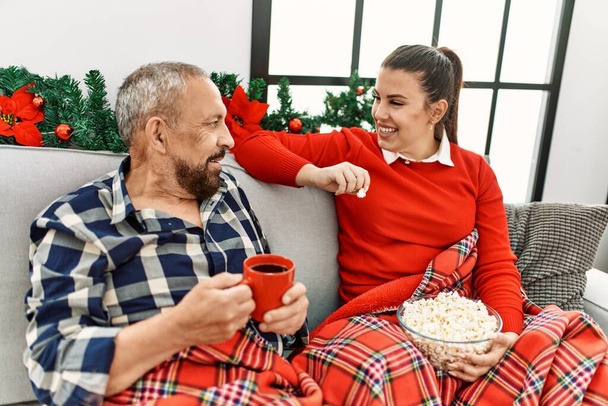 Kleine Tochter und älterer Vater feiern gemeinsam Weihnachten zu Hause, sitzen auf dem Sofa, essen Popcorn und schauen fern - Foto, Bild