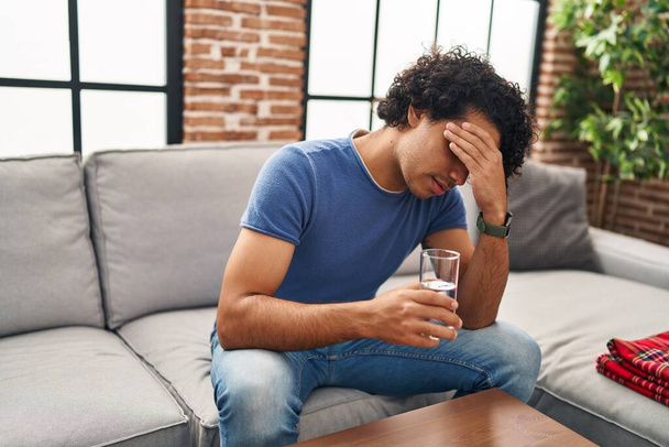 Young hispanic man unwell drinking water sitting on sofa at home - Photo, image
