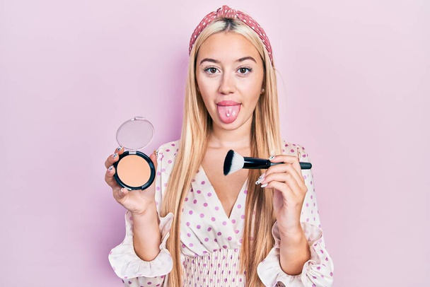 Young blonde girl holding makeup bronzer and brush sticking tongue out happy with funny expression.  - Foto, immagini