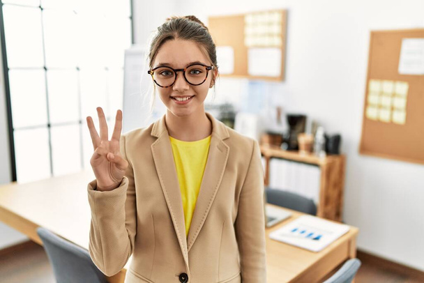 Νεαρή μελαχρινή έφηβη φορώντας επιχειρηματικό στυλ στο γραφείο δείχνει και δείχνει προς τα πάνω με τα δάχτυλα νούμερο τρία, ενώ χαμογελά αυτοπεποίθηση και χαρούμενος.  - Φωτογραφία, εικόνα