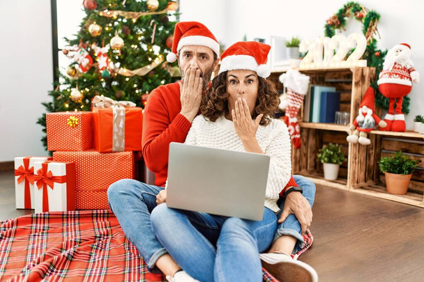 İspanyol orta yaşlı kadın ve olgun erkek dizüstü bilgisayar kullanıyor. Noel ağacının yanında oturup ağzını eliyle kapatıyor. Şok geçiriyor ve hata yapmaktan korkuyor. şaşırmış ifade  - Fotoğraf, Görsel