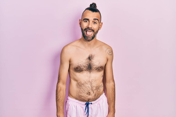 Молодий іспаномовний чоловік без сорочки носить купальник, що стирчить з язиком щасливим виразом сміху. концепція емоцій
.  - Фото, зображення