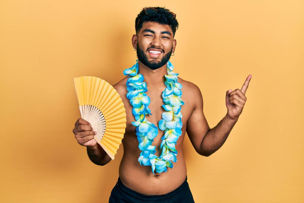 Homem árabe com barba vestindo havaiano lei segurando handfan sorrindo feliz apontando com a mão e o dedo para o lado  - Foto, Imagem