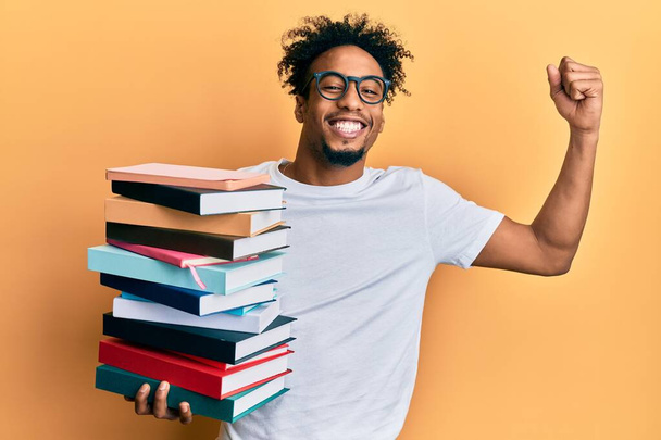 Молодой африканский американец с бородой, держа в руках кучу книг, гордо крича, празднуя победу и успех, очень взволнован поднятой рукой  - Фото, изображение