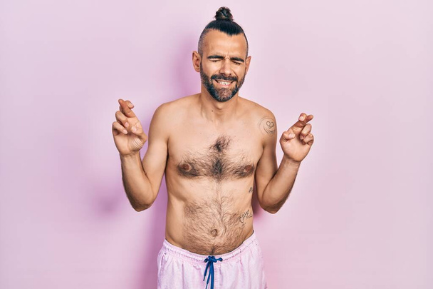 Fiatal spanyol férfi félmeztelenül fürdőruhát visel, ujjal keresztbe tett mosollyal, reménnyel és csukott szemmel. szerencse és babonás fogalom.  - Fotó, kép