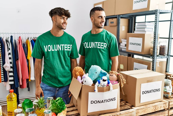 Νεαρό γκέι ζευγάρι που φοράει εθελοντικό μπλουζάκι σε δωρεές στέκεται κοιτάζοντας μακριά στο πλευρό με χαμόγελο στο πρόσωπο, φυσική έκφραση. γελώντας σίγουρος.  - Φωτογραφία, εικόνα