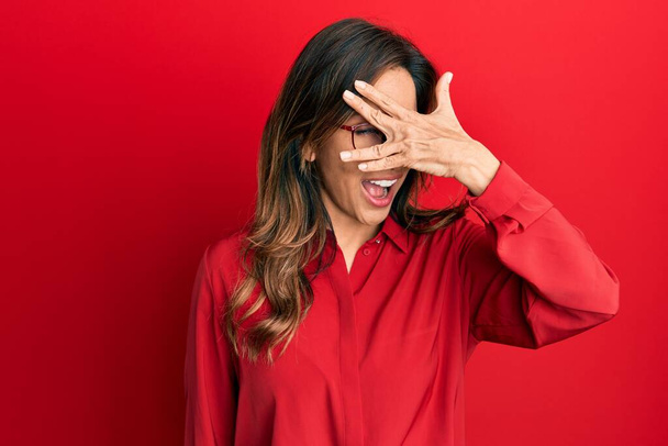 Young latin woman wearing casual clothes and glasses peeking in shock covering face and eyes with hand, looking through fingers with embarrassed expression.  - Foto, afbeelding
