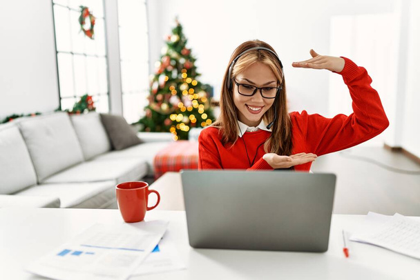 Young caucasian girl sitting on the table working using laptop by christmas tree gesturing with hands showing big and large size sign, measure symbol. smiling looking at the camera. measuring concept.  - Фото, изображение