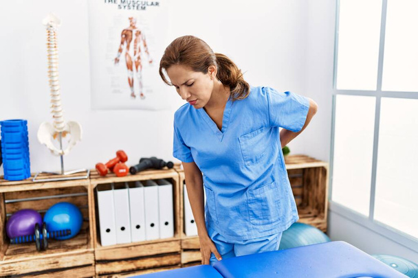 Orta yaşlı İspanyol fizyoterapist kadın ağrı kesici kliniğinde çalışıyor sırt ağrısı çekiyor, elle dokunuyor, kas ağrısı çekiyor.  - Fotoğraf, Görsel