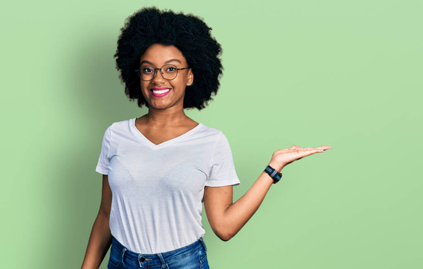 Junge afrikanisch-amerikanische Frau trägt legeres weißes T-Shirt, lächelt fröhlich und zeigt mit der Handfläche in die Kamera.  - Foto, Bild