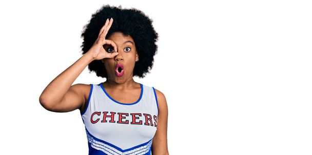 Young african american woman wearing cheerleader uniform doing ok gesture shocked with surprised face, eye looking through fingers. unbelieving expression.  - 写真・画像