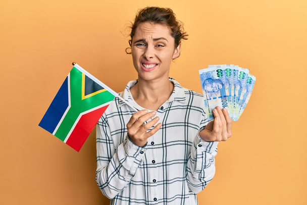 南アフリカの国旗と銀行券を持っている若いブルネットの女性は、無知で混乱した表現。疑わしい概念.  - 写真・画像