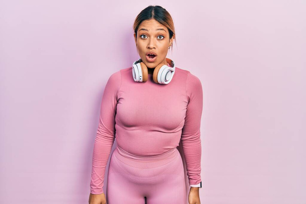 Beautiful hispanic woman wearing gym clothes and using headphones afraid and shocked with surprise and amazed expression, fear and excited face.  - Photo, Image
