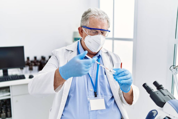 Középkorú ősz hajú férfi, tudományos egyenruhát visel fecskendővel a laboratóriumban. - Fotó, kép