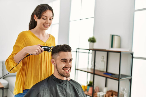 Joven peluquera hispana cortando el pelo del hombre usando máquina de afeitar eléctrica en el centro de belleza. - Foto, imagen