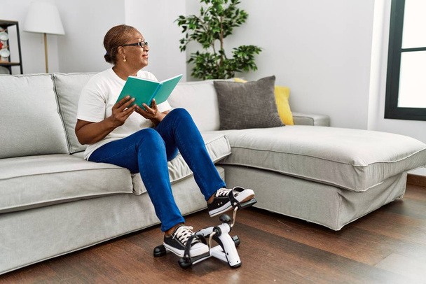 Senior african american woman reading book doing sport using bike pedals machine at home - Photo, Image
