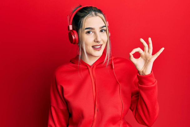 Young modern girl wearing gym clothes and using headphones smiling positive doing ok sign with hand and fingers. successful expression.  - Fotó, kép