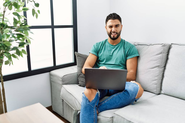 Jeune homme beau avec barbe à l'aide d'un ordinateur portable assis sur le canapé à la maison clin d'oeil en regardant la caméra avec une expression sexy, gai et heureux visage.  - Photo, image