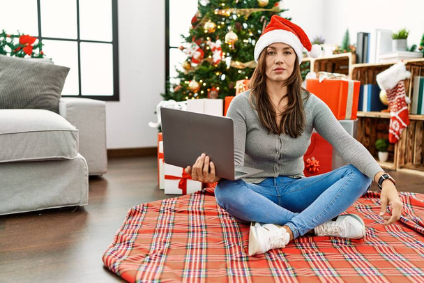 Jeune femme latine utilisant un ordinateur portable assis près de l'arbre de Noël détendu avec une expression sérieuse sur le visage. simple et naturel regardant la caméra.  - Photo, image