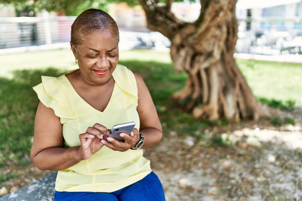 公園のベンチに座っているスマートフォンを使っているシニアアフリカ系アメリカ人女性. - 写真・画像