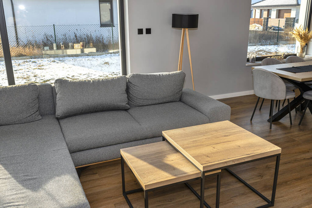 Ein modernes und einfaches Wohnzimmer im Haus mit einem grauen Sofa, sichtbaren großen Terrassenfenstern und zwei Tischen hintereinander. - Foto, Bild