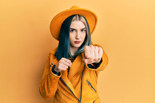 Молодая современная девушка в жёлтой шляпе и кожаной куртке бьет кулаком в драку, агрессивное и злое нападение, угроза и насилие  - Фото, изображение